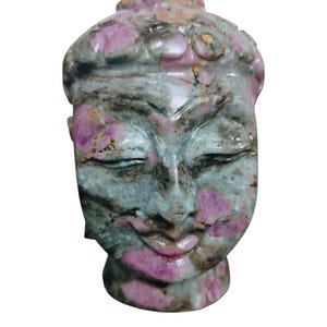 May include: A carved stone Buddha head sculpture. The sculpture features a serene expression with closed eyes and a slight smile. The stone is a mix of gray, pink, and black, with pink accents on the face and head. The sculpture is a spiritual or decorative item.