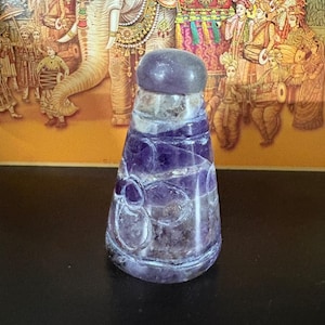 Puede incluir: Un tapón de botella de amatista púrpura con una parte superior redondeada y un diseño tallado. El tapón mide aproximadamente 7,6 cm de alto. El fondo presenta una colorida ilustración de un elefante y personas con vestimenta tradicional.