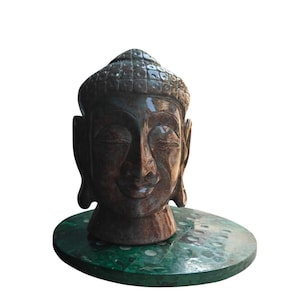 Peut inclure: Une sculpture détaillée d'une tête de Bouddha, avec une expression sereine et les yeux fermés. La tête est de couleur marron foncé et repose sur une base ronde verte. La sculpture est probablement en pierre ou dans un matériau similaire.