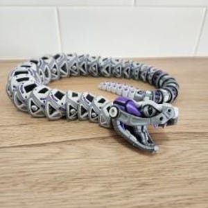 May include: A 3D-printed articulated snake toy in shades of grey, white, and purple. The snake's body is segmented, allowing for flexible posing. The head features detailed eyes and an open mouth, showcasing the intricate design. The toy is approximately 30 cm long.