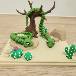 May include: A whimsical desert scene featuring a green snake winding around a brown tree and two cacti. The scene includes a beige base with a light brown background and two green snakes with cartoonish eyes. The cacti are green with white dots.