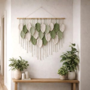 Könnte beinhalten: Makramee-Wandbehang mit Blattdesign in Grün- und Off-White-Tönen. Die Blätter sind an einer natürlichen Holzstange mit langen, herabhängenden Fransen befestigt. Die Wanddekoration wird vor einer weißen Wand präsentiert.