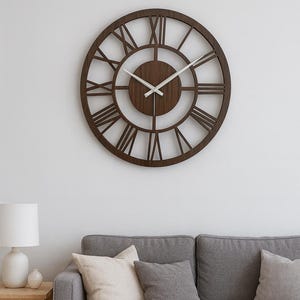 Puede incluir: Un reloj de pared redondo de madera con números romanos. El reloj es marrón con manecillas blancas y está montado en una pared blanca. Debajo, un sofá gris con cojines. Una lámpara y una mesa auxiliar a la izquierda.
