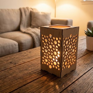 Könnte beinhalten: Eine Holzlaterne mit geometrischem Muster, von innen beleuchtet, steht auf einem rustikalen Holztisch. Das warme Licht scheint durch das ausgeschnittene Design. Eine Sukkulente in einem weißen Topf ist im Hintergrund zu sehen.