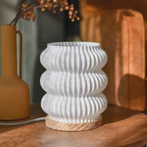 Könnte beinhalten: Eine weiße Tischlampe mit Rippen und einem Holzsockel. Die Lampe hat ein einzigartiges, gestapeltes Design und ist an eine Steckdose angeschlossen. Eine gelbe Vase und dekorative Zweige befinden sich im Hintergrund und schaffen eine warme Atmosphäre.