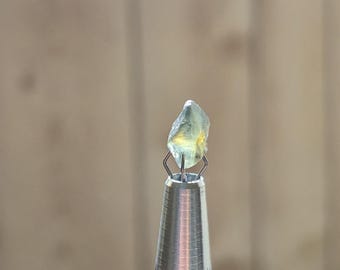 May include: A raw, pale yellow-green gemstone is held by a silver-coloured tool. The gemstone is irregularly shaped with a slightly translucent appearance. The tool has a textured, metallic handle and thin wire prongs to secure the stone.