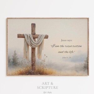 Può includere: Una stampa artistica rettangolare da parete raffigurante una croce in legno drappeggiata con un panno beige chiaro, su un paesaggio nebbioso con alberi ed erba. Il testo recita: Jesus says: I am the resurrection and the life. John 11, 25. Art & Scripture by Ina.