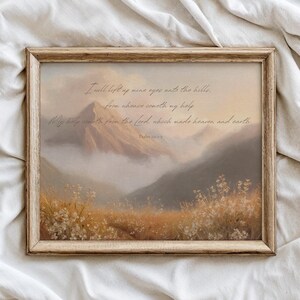 Könnte beinhalten: Gerahmter Kunstdruck mit einer Berglandschaft und einem Feld mit Wildblumen. Das Kunstwerk hat eine warme Farbpalette und enthält Text aus Psalm 121:1-2. Der Rahmen ist hellbraun.