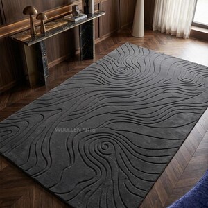 May include: A rectangular dark gray area rug featuring a carved, raised relief pattern of swirling, organic lines that resemble topographical contours or wood grain. The text WOLLEN ARTS is visible in a subtle font on the rug surface.