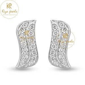 May include: A pair of silver-toned stud earrings. Each earring has a curved, wing-like design, encrusted with numerous small, sparkling white gemstones. The earrings are set against a white background, with the "Kiza Jewels" logo visible.