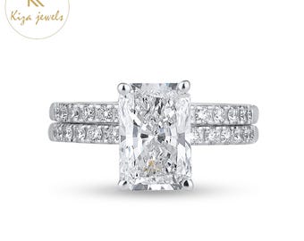 Puede incluir: Un anillo de compromiso con diamantes de talla radiante y una banda a juego. La piedra principal es un diamante rectangular grande engastado en una banda plateada, flanqueado por diamantes más pequeños. El logotipo "Kiza jewels" está en la esquina superior izquierda.