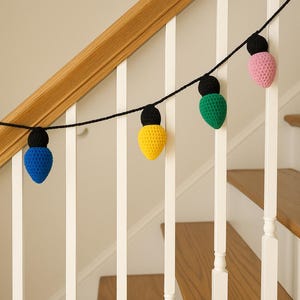 Może przedstawiać: Girlanda kolorowych, szydełkowych ozdób w kształcie żarówek w kolorze niebieskim, żółtym, zielonym i różowym, każda z czarną podstawą. Lampki są nawleczone na czarny sznurek, zawieszone na poręczy schodów.