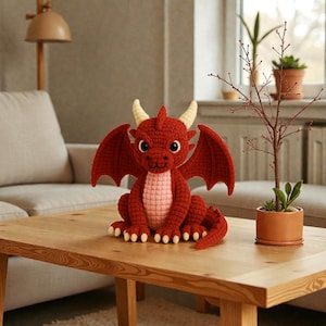 May include: A handmade red crochet dragon with cream-colored horns, large wings, and a light pink belly sits on a wooden table. The dragon has large, round eyes and is a whimsical decorative item.