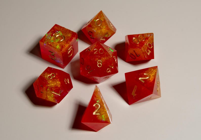 May include: Set of seven polyhedral dice in various shapes. The dice are a translucent red with gold and yellow internal marbling. Each die has gold numbering. The dice are arranged on a white surface.