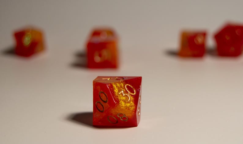 May include: A set of polyhedral dice with a red and gold color scheme. The dice are made of a translucent material, with gold-colored numbers. The dice are designed for tabletop role-playing games.