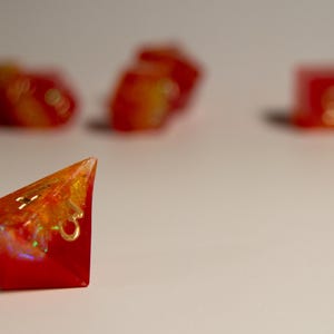 May include: Red and orange resin dice set. The image shows a single, triangular die in sharp focus, with others blurred in the background. The dice have a vibrant, translucent appearance with gold accents, suggesting a fantasy game theme.