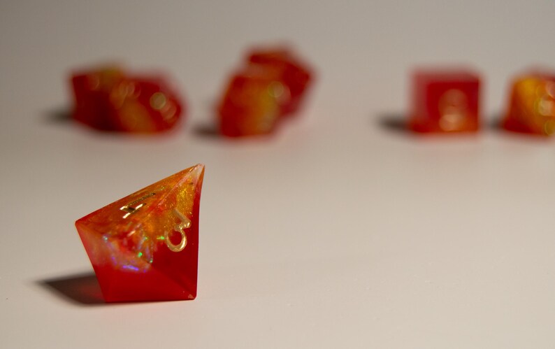 May include: Red and orange resin dice set. The image shows a single, triangular die in sharp focus, with others blurred in the background. The dice have a vibrant, translucent appearance with gold accents, suggesting a fantasy game theme.