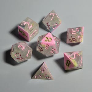 Può includere: Un set di sette dadi poliedrici di varie forme. I dadi sono trasparenti con vortici rosa e verdi e numeri rosa. Questi dadi sono progettati per giochi di ruolo da tavolo.