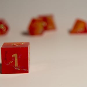 May include: A red resin die with the number 1 in gold. The die has a glittery, marbled appearance with orange and yellow accents. Other dice are blurred in the background.
