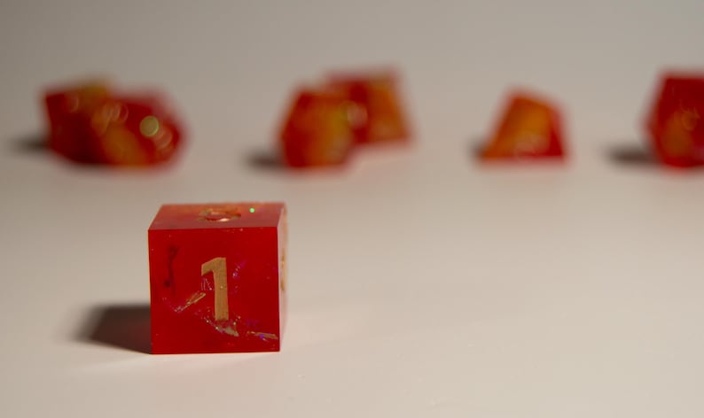 May include: A red resin die with the number 1 in gold. The die has a glittery, marbled appearance with orange and yellow accents. Other dice are blurred in the background.