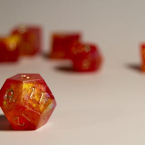 May include: A set of polyhedral dice in shades of red and gold. The twelve-sided die in the foreground is in sharp focus, displaying the number 12. Other dice are blurred in the background, suggesting a game or collection.