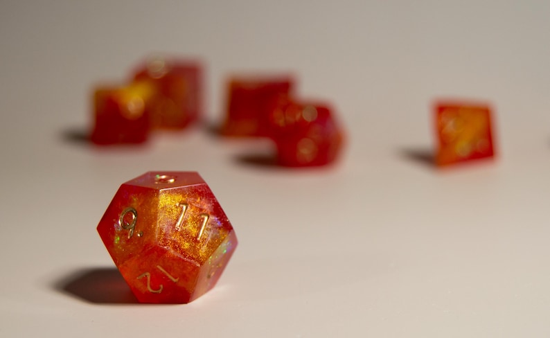May include: A set of polyhedral dice in shades of red and gold. The twelve-sided die in the foreground is in sharp focus, displaying the number 12. Other dice are blurred in the background, suggesting a game or collection.