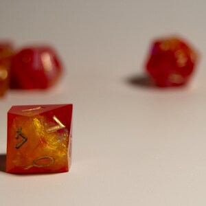 May include: A collection of handmade, resin dice in various shapes. The dice are primarily red with gold and orange inclusions, giving them a fiery appearance. The dice are designed for tabletop games.