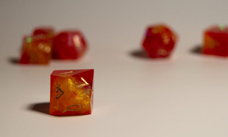 May include: A collection of handmade, resin dice in various shapes. The dice are primarily red with gold and orange inclusions, giving them a fiery appearance. The dice are designed for tabletop games.