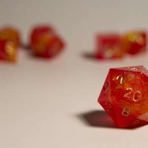 May include: A red and orange twenty-sided die, with gold numbering, is in focus. Several other dice are out of focus in the background. The dice have a fiery, translucent appearance, suggesting a fantasy game theme.