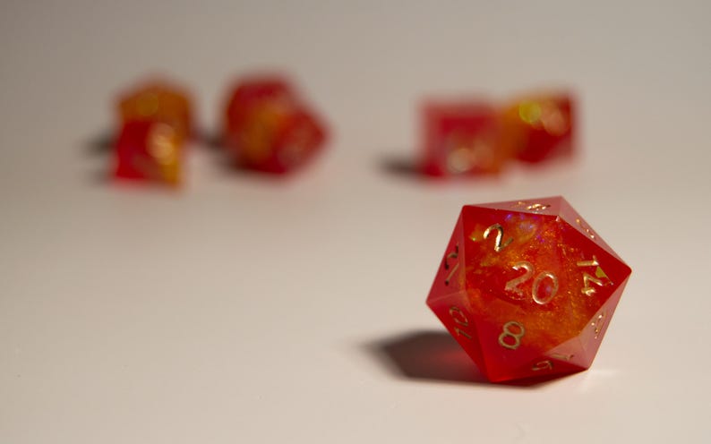 May include: A red and orange twenty-sided die, with gold numbering, is in focus. Several other dice are out of focus in the background. The dice have a fiery, translucent appearance, suggesting a fantasy game theme.