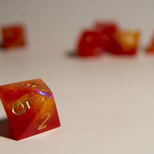 May include: Close-up of a polyhedral die with a red and orange color scheme. The die has gold-colored numbers and is made of a translucent material. Other dice are out of focus in the background.
