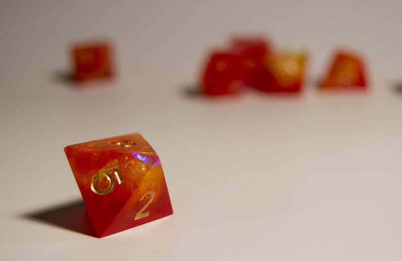 May include: Close-up of a polyhedral die with a red and orange color scheme. The die has gold-colored numbers and is made of a translucent material. Other dice are out of focus in the background.