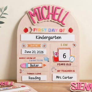 Könnte beinhalten: Ein helles, bogenförmiges Holzschild mit dem Namen MICHELL in Pink, unterteilt in Bereiche für First Day Of, Kindergarten, Datum 20. Juni 2026, Alter 6, Berufswunsch Baker und Lehrerin Mrs. Carter, inklusive zusätzlicher Namensschilder und Etiketten.