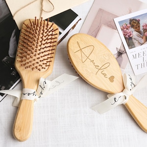 Könnte beinhalten: Zwei Holzbürsten mit hellbraunen Borsten und Griffen. Eine ist rechteckig, die andere oval, beide mit dem eingravierten Namen "Amelia". Weißes Band mit Schreibschrifttext ist um die Griffe gebunden. Eine Karte mit dem Text "Meet You Like The Wine" befindet sich im Hintergrund.