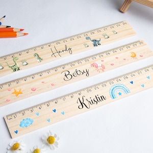 Puede incluir: Tres reglas de madera, cada una de 50 cm de largo, con nombres personalizados e ilustraciones decorativas. Una regla tiene un tema de dinosaurios, otra de unicornios y la tercera de arcoíris. Cada regla tiene marcas en pulgadas y centímetros.