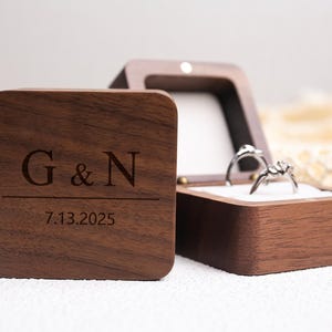 Könnte beinhalten: Eine quadratische, dunkle Holzringbox mit den eingravierten Initialen "G & N" und dem Datum "7.13.2025". Die Box ist geöffnet und zeigt zwei Ringe. Die Ringe sind silberfarben mit einem floralen Design. Die Box steht neben einer passenden geschlossenen Box.