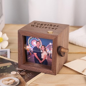Könnte beinhalten: Ein hölzerner Fotowürfel mit einem Foto eines Paares vor einem Riesenrad. Auf dem Würfel steht "Together Wherever". Oben befindet sich ein kleiner, goldfarbener Akzent und an der Seite ein Holzknopf. Das Holz ist dunkel.