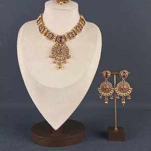 Peut inclure: Un ensemble collier et boucles d'oreilles doré orné de motifs circulaires avec des pierres rouge foncé incrustées. Le collier présente un pendentif central avec des perles pendantes, et les boucles d'oreilles sont circulaires avec des accents similaires.