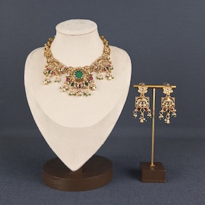 Peut inclure: Un ensemble collier et boucles d'oreilles pendantes doré avec un travail du métal complexe, une pierre verte centrale sur le collier, et des accents de petites pierres rouges et vertes avec des perles blanches pendantes sur les bijoux.