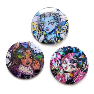 Op de afbeelding: Drie ronde holografische knoppen met cartoonfiguren op kleurrijke achtergronden. De eerste knop heeft een blauwhuidige figuur met groen haar en een bliksem. De tweede knop heeft een bruinhuidige figuur met lang bruin haar en een halve maan. De derde knop heeft een rozehuidige figuur met roze haar en een zwarte hoed.