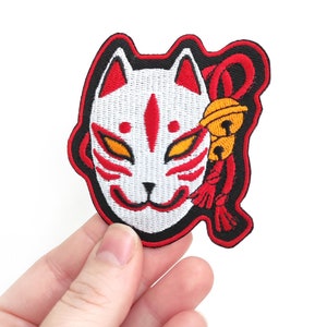 May include: White and red embroidered iron-on patch of a Japanese kitsune mask with a red and gold bell hanging from a black cord.