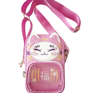 May include: Pink crossbody bag with a cat face design. The bag features a cat face with embroidered details and a clear pocket with Japanese text. It has a pink adjustable strap and gold-coloured hardware.