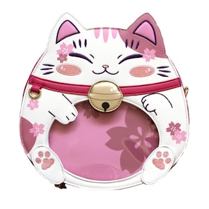 Puede incluir: Bolso con forma de gato blanco y rosa con ventana transparente. El bolso presenta una cara de gato sonriente, detalles de flores de cerezo rosas y un collar rosa con una campana dorada. El interior es rosa y tiene un diseño de huella de pata.