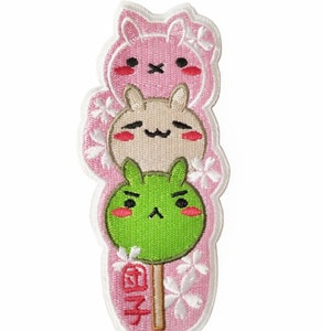 May include: Embroidered patch featuring three cartoon bunny-shaped dango on a stick. The top bunny is pink, the middle is tan, and the bottom is green. The patch has a white border and pink background with white flower accents. Japanese text is visible.