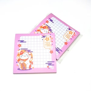 Puede incluir: Notas adhesivas cuadradas rosas con un patrón de cuadrícula y una ilustración de gato de la suerte japonés. El gato es blanco con detalles en naranja y rojo, sosteniendo una moneda de oro. Las notas también presentan texto japonés y diseños florales.