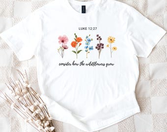 Könnte beinhalten: Weißes T-Shirt mit einem Aquarell-Blumenmuster. Der Text "LUKE 12:27" steht über den Blumen und "consider how the wildflowers grow" darunter. Die Blumen sind in Rosa, Orange, Blau, Lila und Gelb gehalten.
