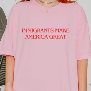 Może przedstawiać: Jasnoróżowa koszulka z czerwonym napisem "IMMIGRANTS MAKE AMERICA GREAT". Koszulka ma krótkie rękawy i luźny krój. Tekst jest umieszczony centralnie z przodu.