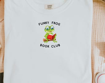 Może przedstawiać: Biała koszulka z okrągłym dekoltem z grafiką zielonej żaby w okularach czytającej czerwoną książkę. Tekst "FUNKY FROG BOOK CLUB" znajduje się nad i pod ilustracją żaby.