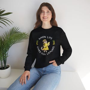 Könnte beinhalten: Schwarzes Sweatshirt mit dem Text "WHEN LIFE GIVES YOU LEMONS" und einem Cartoon-Husky in gelbem Regenmantel, der Zitronen hält. Das Sweatshirt hat einen Rundhalsausschnitt.