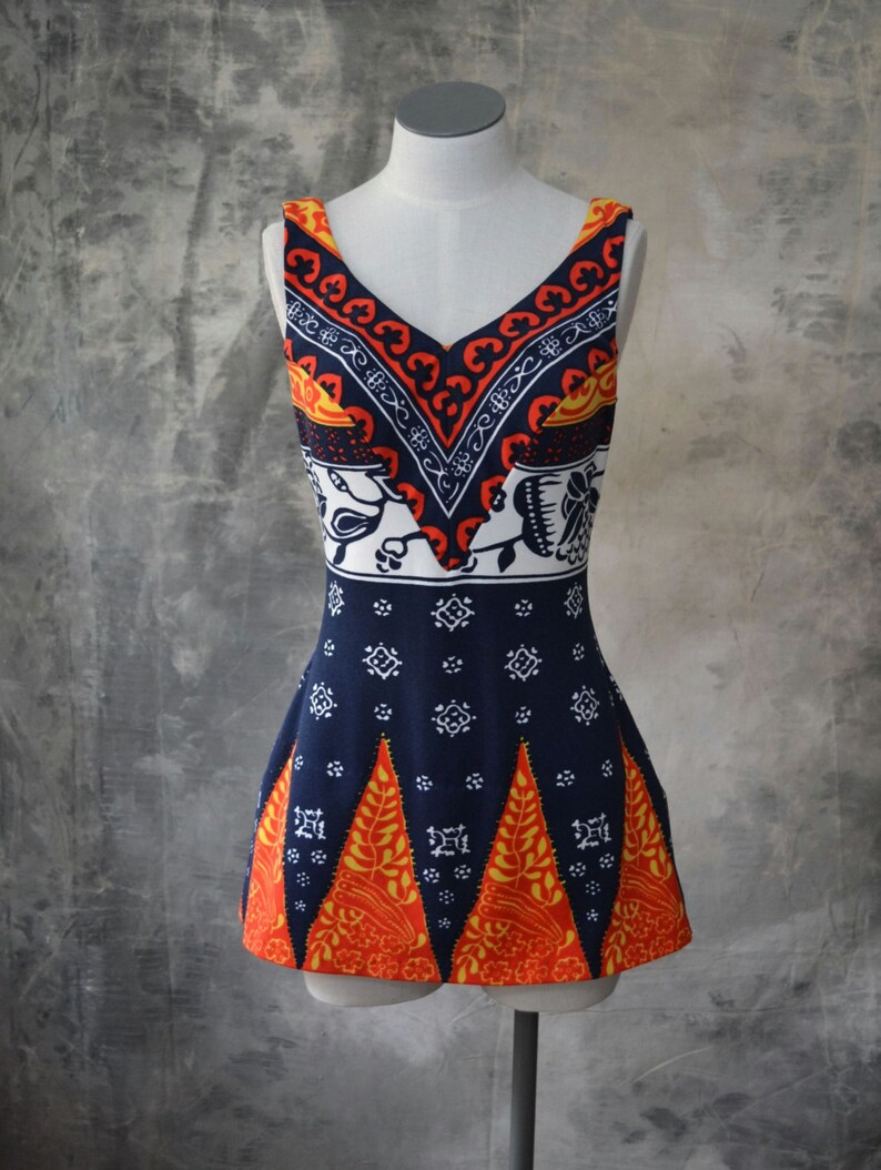 May include: A sleeveless, V-neck top on a mannequin. The top has a navy blue base with white, orange, and yellow geometric and floral patterns. The top has a fitted bodice and a flared hemline, showcasing a vintage style.